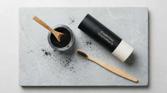A minimalist flat-lay of charcoal toothpaste, charcoal powder in an open jar, and a bamboo toothbrush arranged on a stone surface with soft natural lighting.