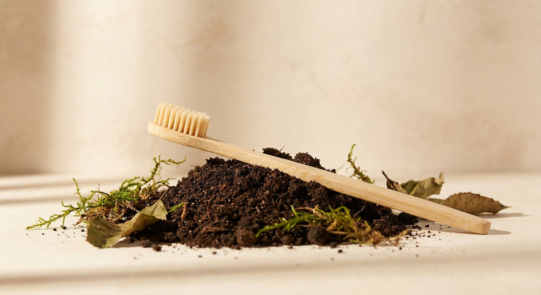 A bamboo toothbrush resting on dark compost soil surrounded by natural dried leaves and moss, evoking biodegradability and the return to earth.
