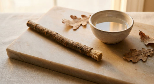 A fresh miswak stick resting on a natural stone surface beside a small ceramic bowl of water, styled with dried leaves in warm earthy tones.