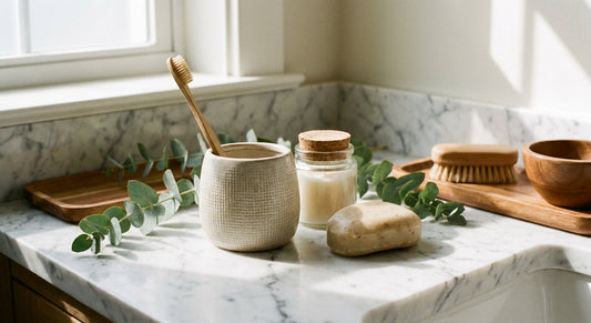 A sunlit bathroom countertop featuring a bamboo toothbrush, glass jar, natural soap bar, and eucalyptus sprigs arranged on a clean stone surface.