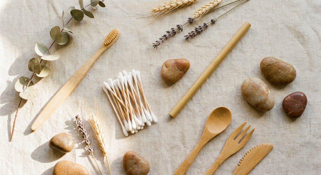 A flat lay of assorted bamboo everyday items including a toothbrush, cotton swabs, straw, and utensils arranged on a natural linen surface with dried botanicals and stones.