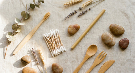 A flat lay of assorted bamboo everyday items including a toothbrush, cotton swabs, straw, and utensils arranged on a natural linen surface with dried botanicals and stones.