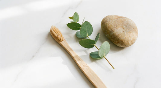 A bamboo toothbrush resting on a white marble surface beside a eucalyptus sprig and smooth stone in soft natural light.